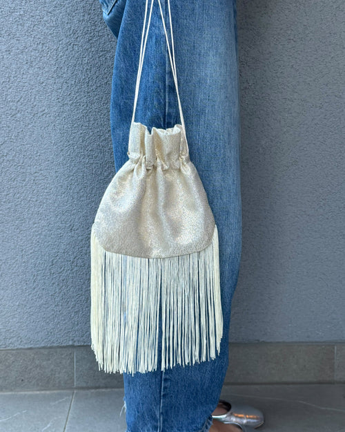 A cream-colored fringe drawstring bag with a metallic gold top panel, hanging from a person's waist over blue jeans.