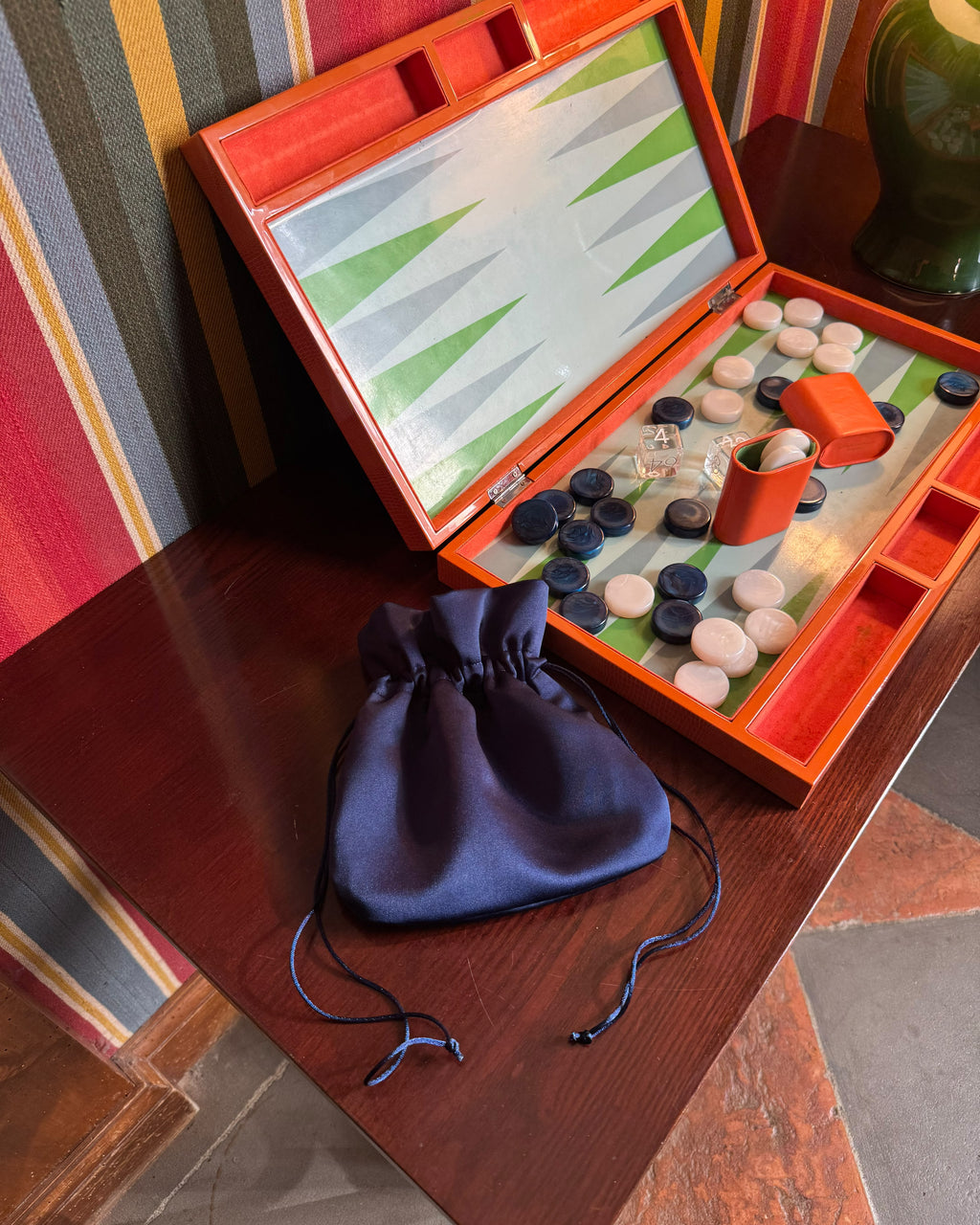 An open wooden backgammon set with a green and white board, orange-red frame, checkers, dice cup, and a navy blue drawstring pouch placed on a dark wooden table. Striped wallpaper and part of a green ceramic vase are visible in the background.