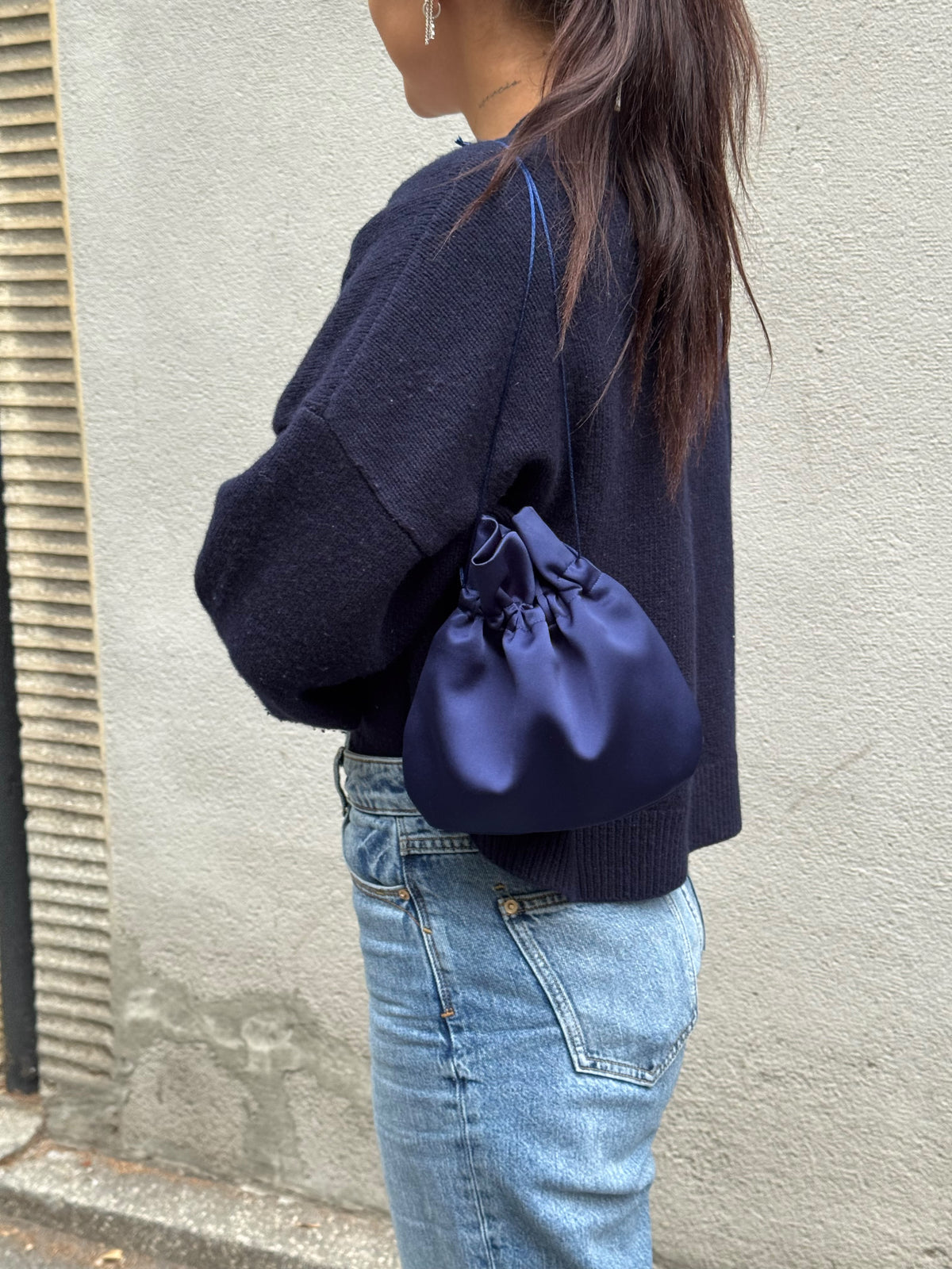 A person wearing a dark navy knit sweater and light blue jeans carries a navy blue satin drawstring pouch bag against a textured gray wall.