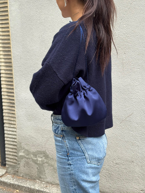 A person wearing a dark navy knit sweater and light blue jeans carries a navy blue satin drawstring pouch bag against a textured gray wall.