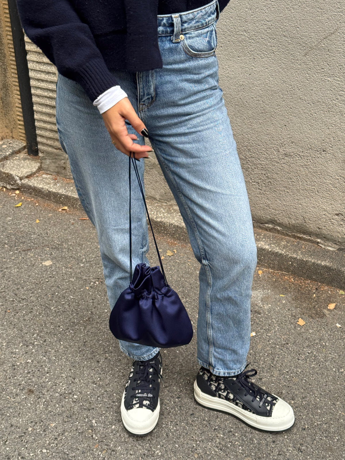 A person wearing light-wash blue jeans, a dark navy sweater, and black-and-white sneakers holds a small navy satin drawstring pouch bag while standing outdoors on a paved street next to a concrete wall.