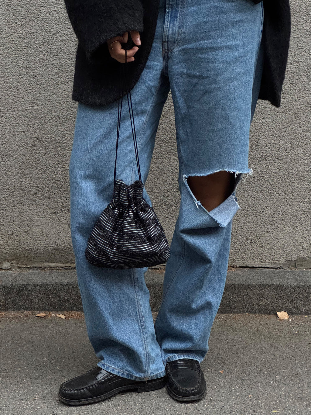A person wearing light-wash blue ripped jeans, black shoes, and a black fuzzy top, holding a black and white striped drawstring pouch bag against a gray concrete wall.