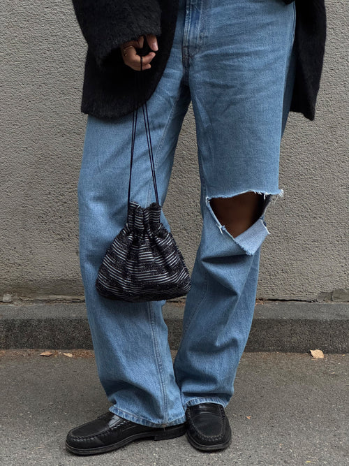 A person wearing light-wash blue ripped jeans, black shoes, and a black fuzzy top, holding a black and white striped drawstring pouch bag against a gray concrete wall.