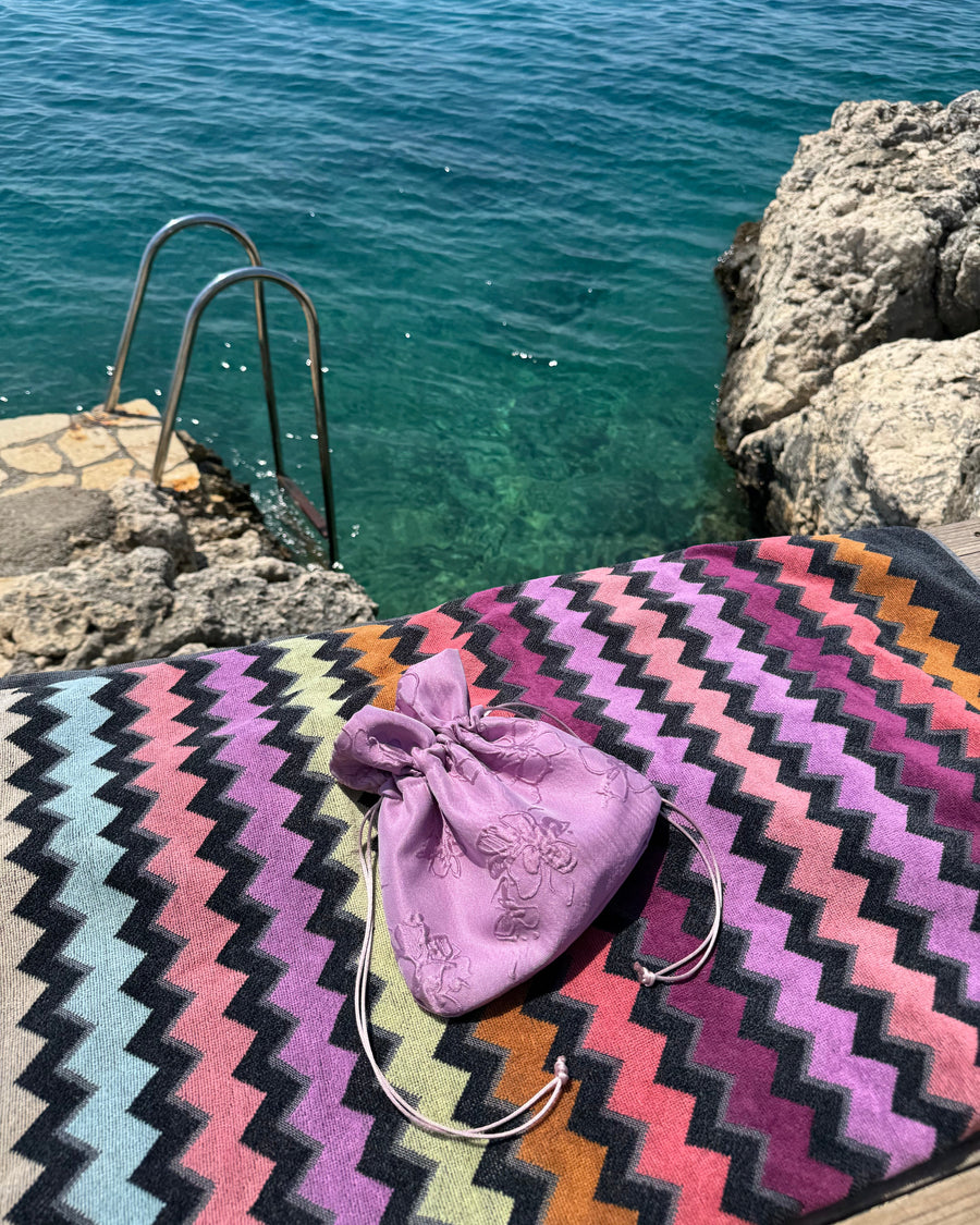 A lavender embroidered drawstring pouch bag resting on a colorful zigzag-patterned towel by the ocean.