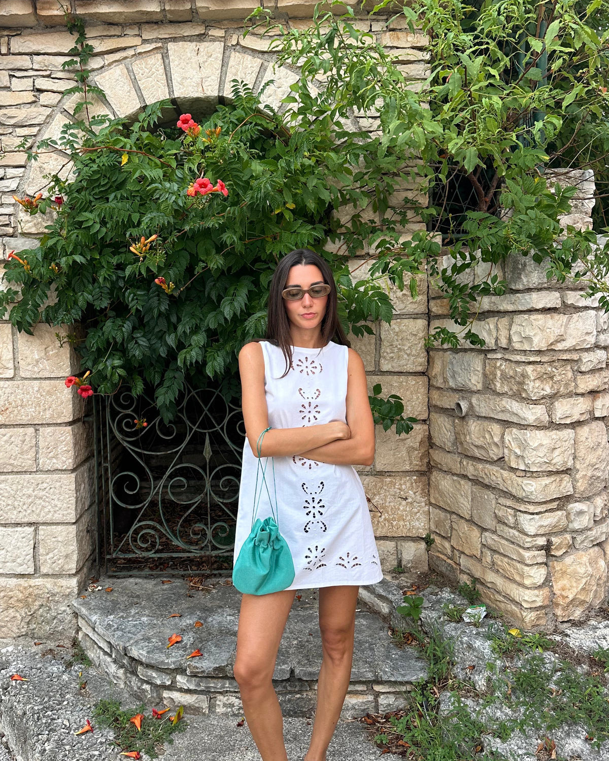 A woman stands outdoors in front of a stone archway with greenery and red flowers, wearing a white sleeveless eyelet dress, sunglasses, and carrying a turquoise shoulder bag.