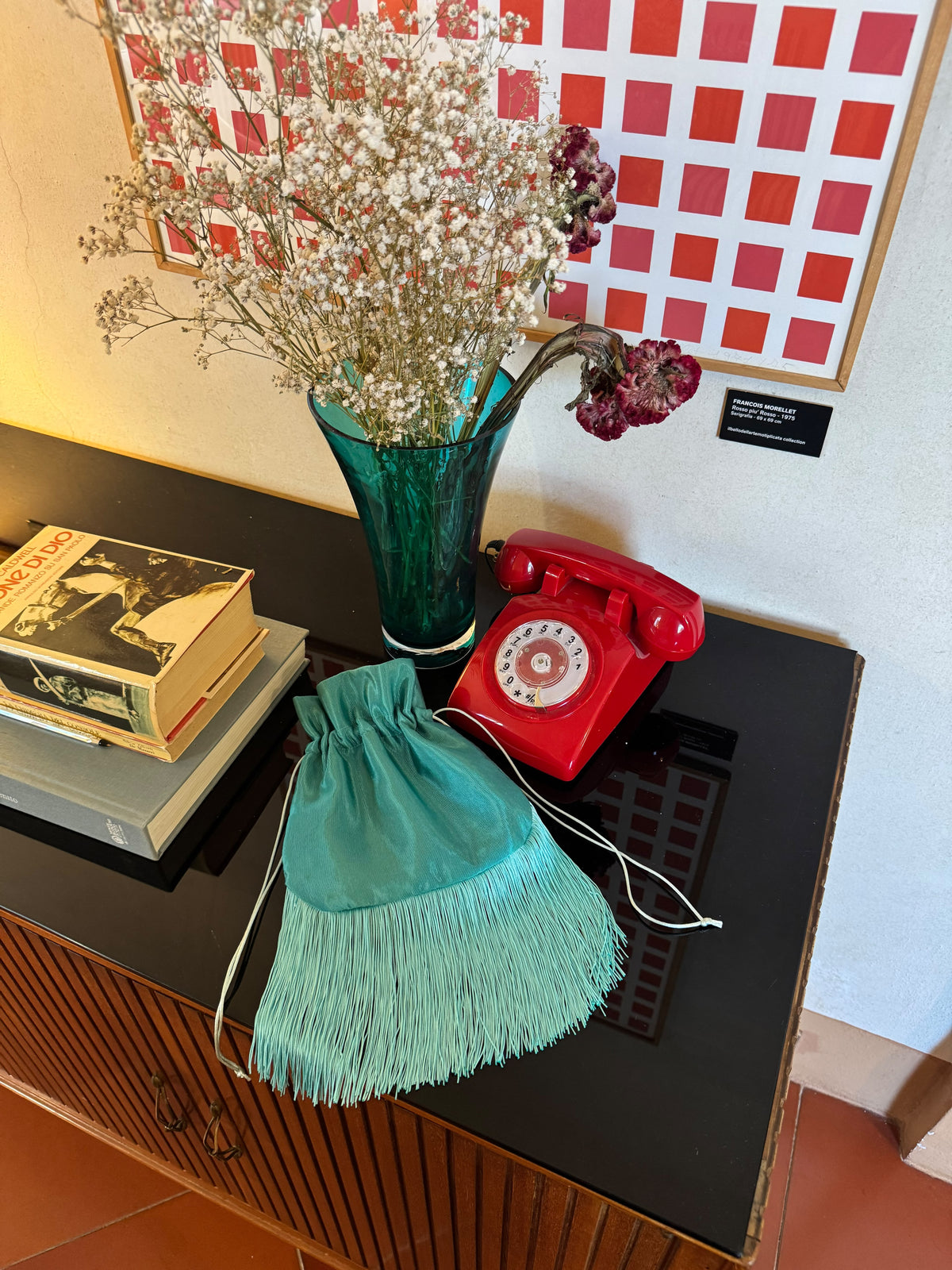 A mint green fringed drawstring pouch displayed on a black wooden cabinet next to a red rotary telephone, a teal glass vase with dried flowers, and books, with a red and white checkered wall art piece in the background.