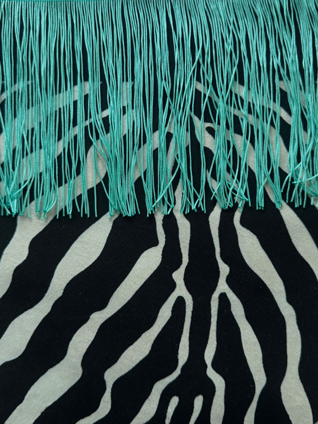 A close-up of a textile featuring a black and off-white zebra stripe pattern with bright turquoise fringe along the top edge.